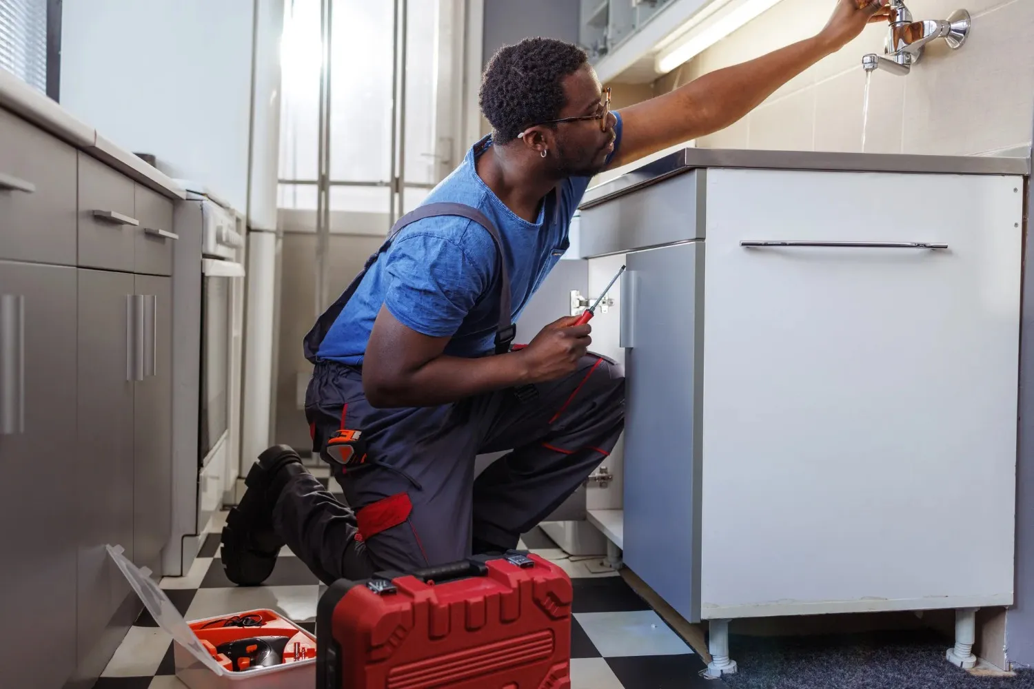  A Black male plumber in blue overalls and a blue shirt is kneeling on a checkered floor, using a screwdriver to work under a white kitchen cabinet, with a red toolbox open beside him.