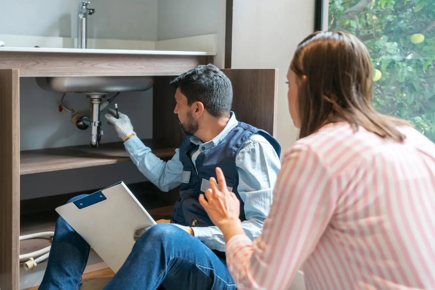  A female customer with long brown hair is watching a male plumber in a blue vest and light blue shirt, wearing white gloves, work on pipes under a sink in a wooden cabinet. He is holding a clipboard.