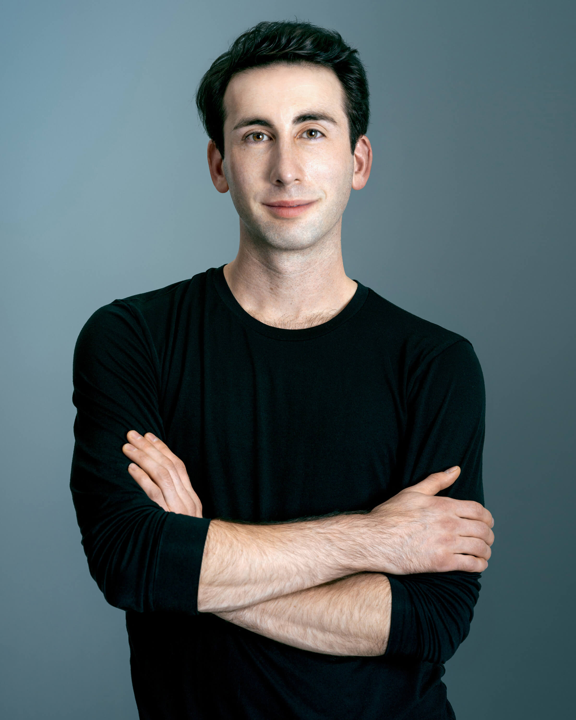 Jacob Kemp wearing a black sweater, standing against a neutral backdrop with his arms crossed, a knowing smile across his face. 