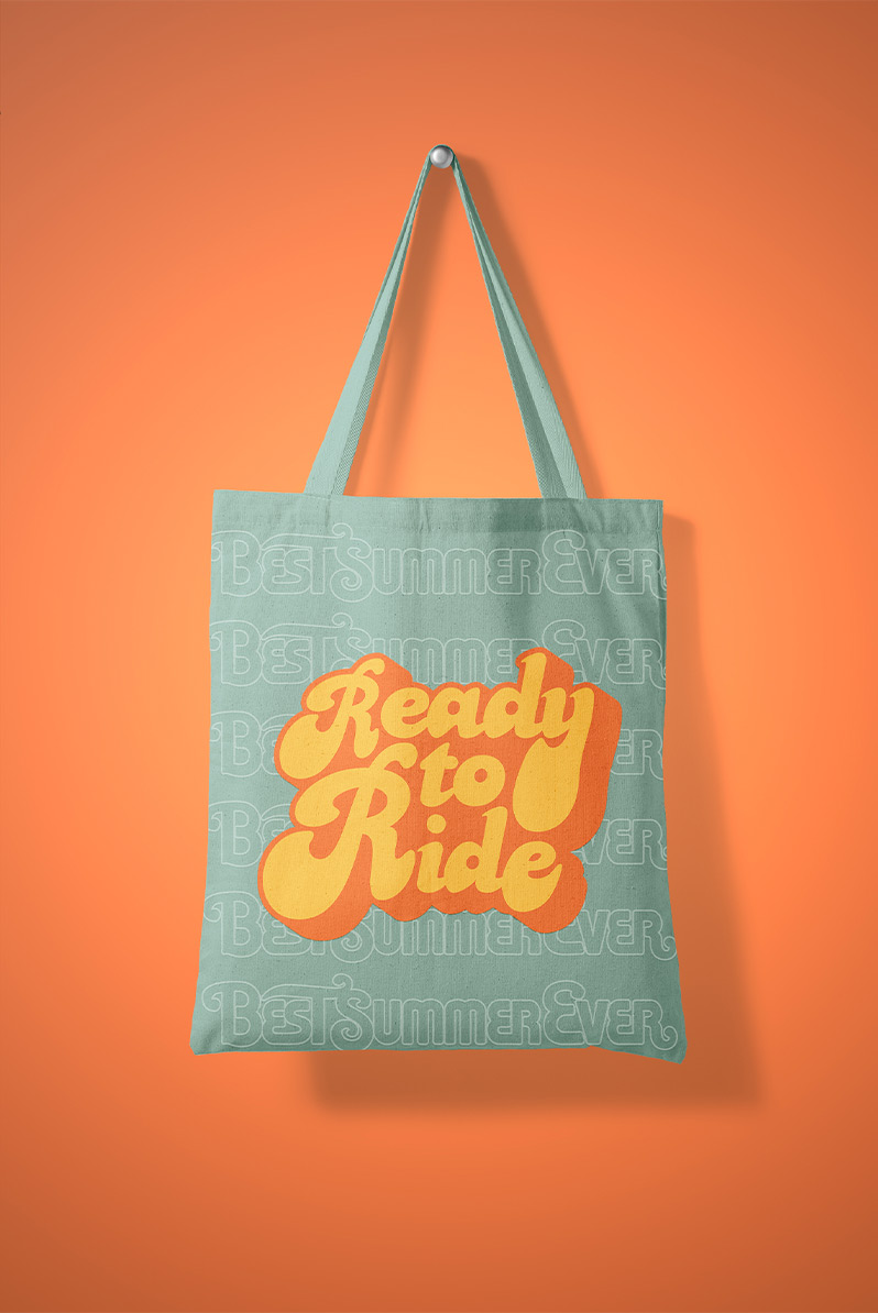 This is an image of a blue-colored tote bag on an orange backdrop. The tote reads "ready to ride" in an orange, hand-lettered font that feels like it was made in the 70s.