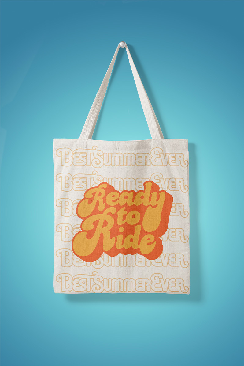 This is an image of a cream-colored tote bag on a blue backdrop. The tote reads "ready to ride" in an orange, hand-lettered font that feels like it was made in the 70s.