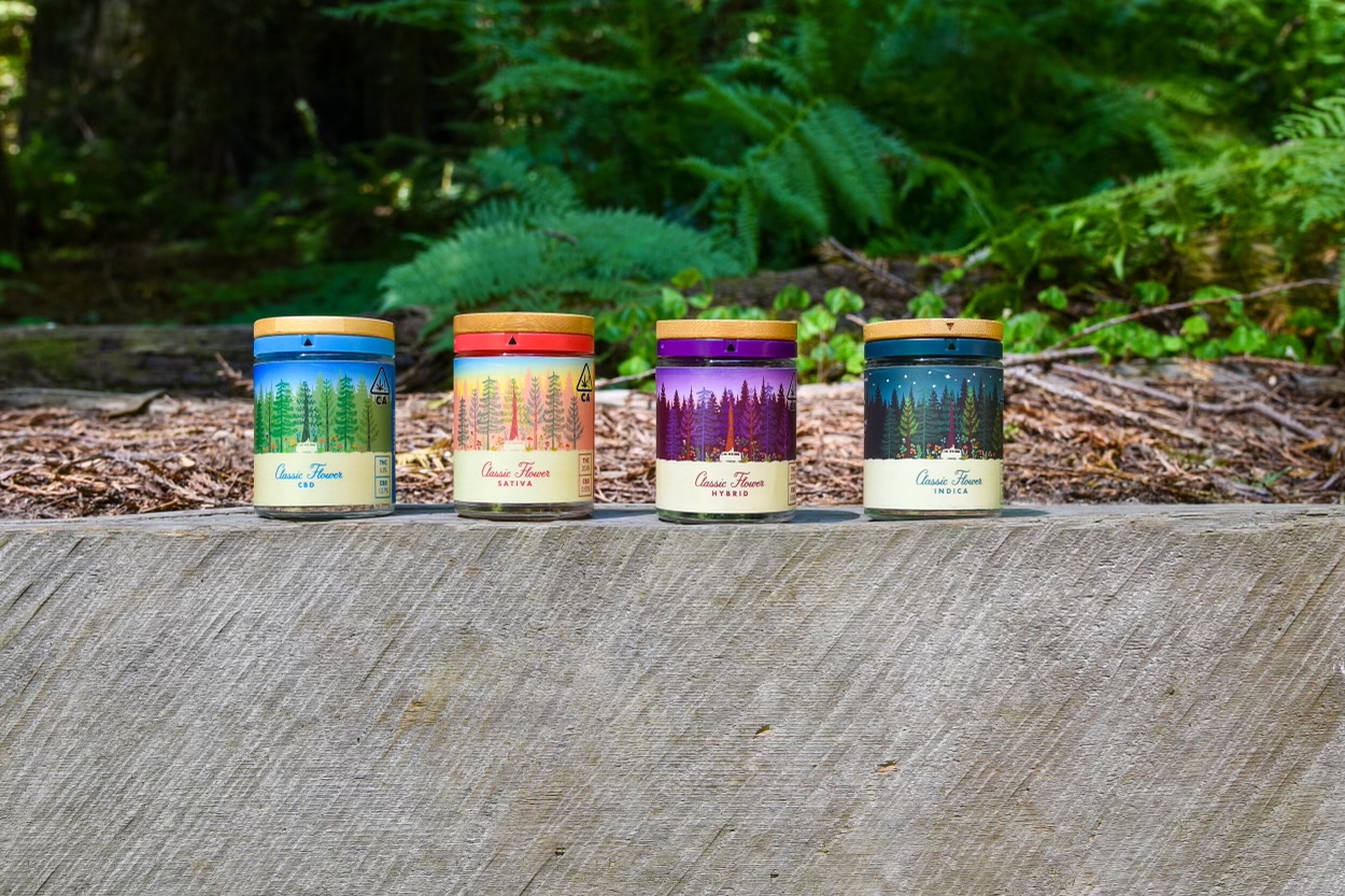 This is an image of four different containers containing loose marijuana flower from Humboldt Farms, photographed on a concrete ledge with a forest in the background. The color of the containers is different, depending on the varietal it contains. The red package contains Sativa, the dark green one contains Indica, the light blue one contains CBD, and the purple one contains a hybrid variety. 