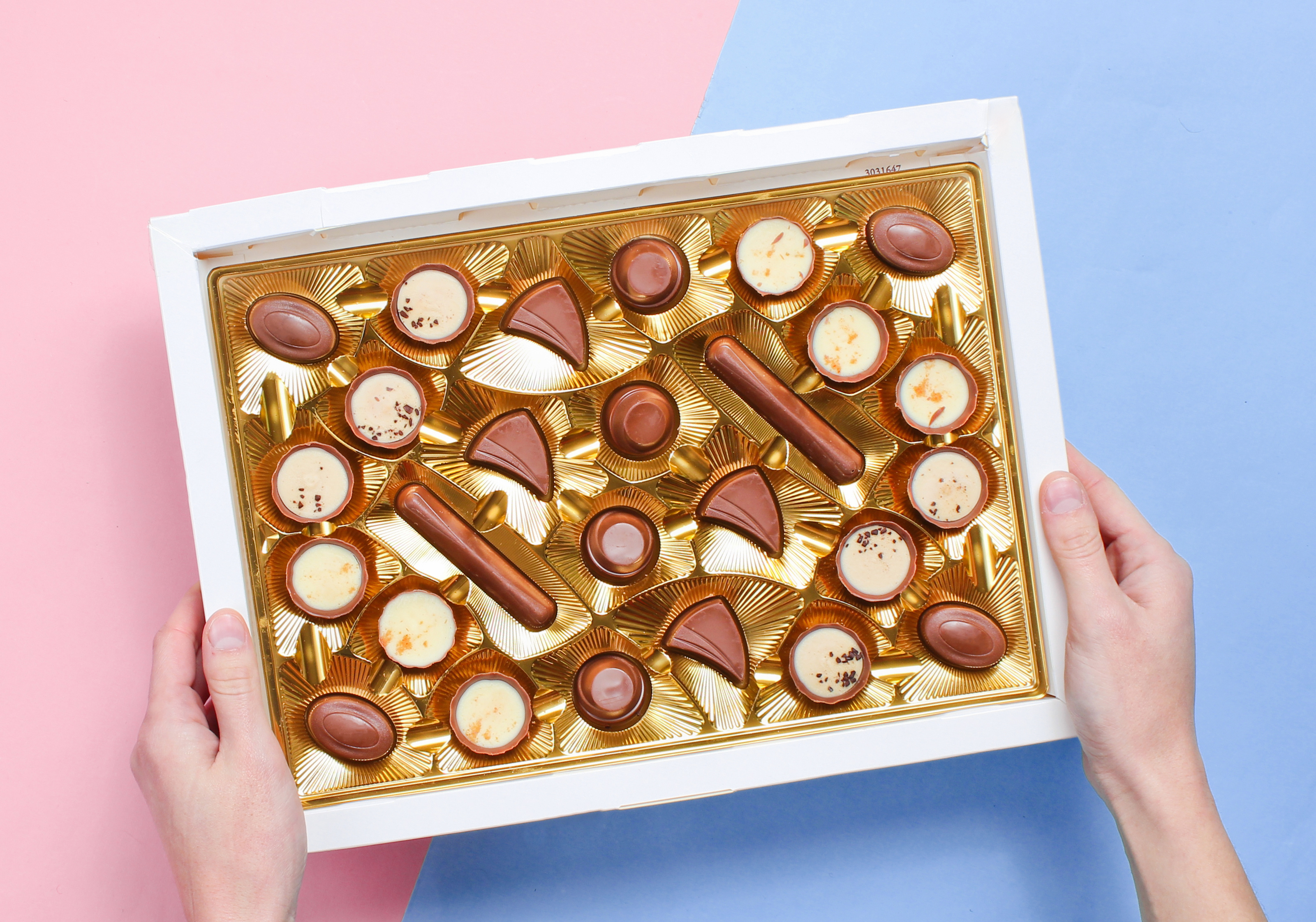 This is an image of a case of 20+ truffles from Chocolaterie Bob. They come in a variety of sizes and types, and are surrounded by a gold foil case. The background is blue and pink, and the image feels premium and cheerful.