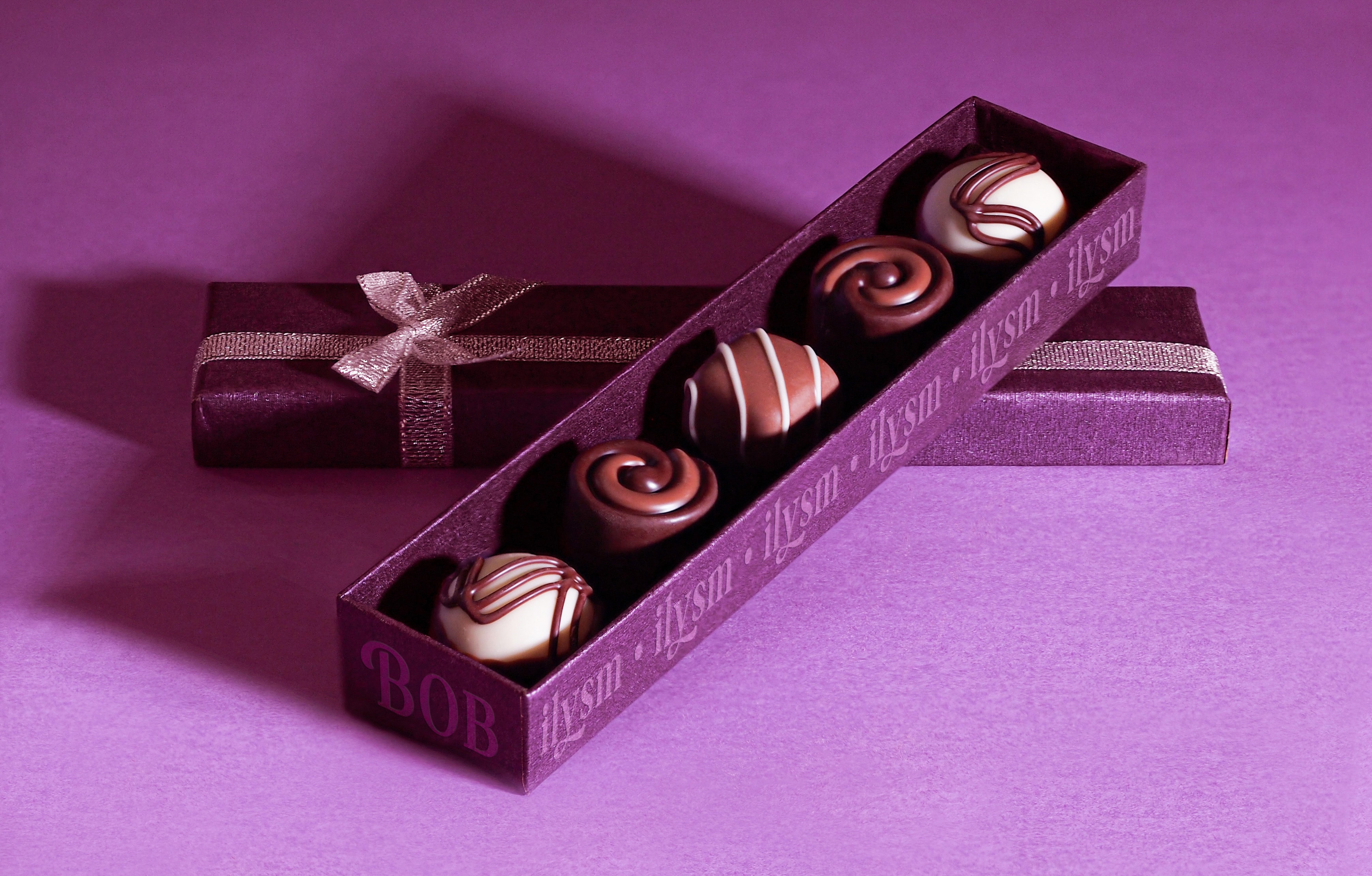 This is a photograph of a case of 5 chocolate truffles from Chocolaterie Bob. The box is a rich purple and the top is framed by a bow. The background is purple and the packaging feels luxurious but playful, because acronyms line the side of the bottom half of the box.;