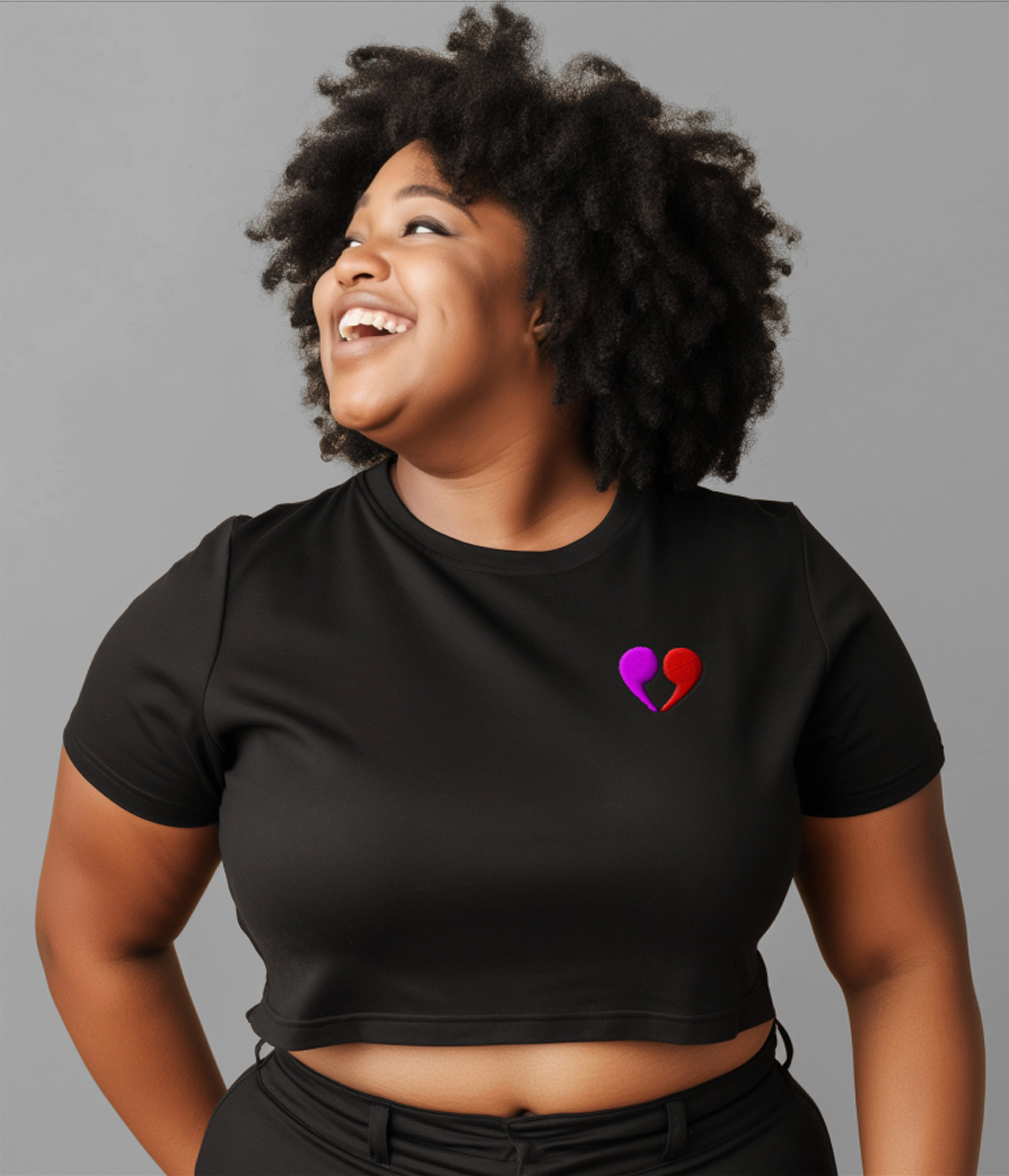 This is an image of a woman with an afro wearing a black crop top with the heart quotation symbol embroidered over her heart. 