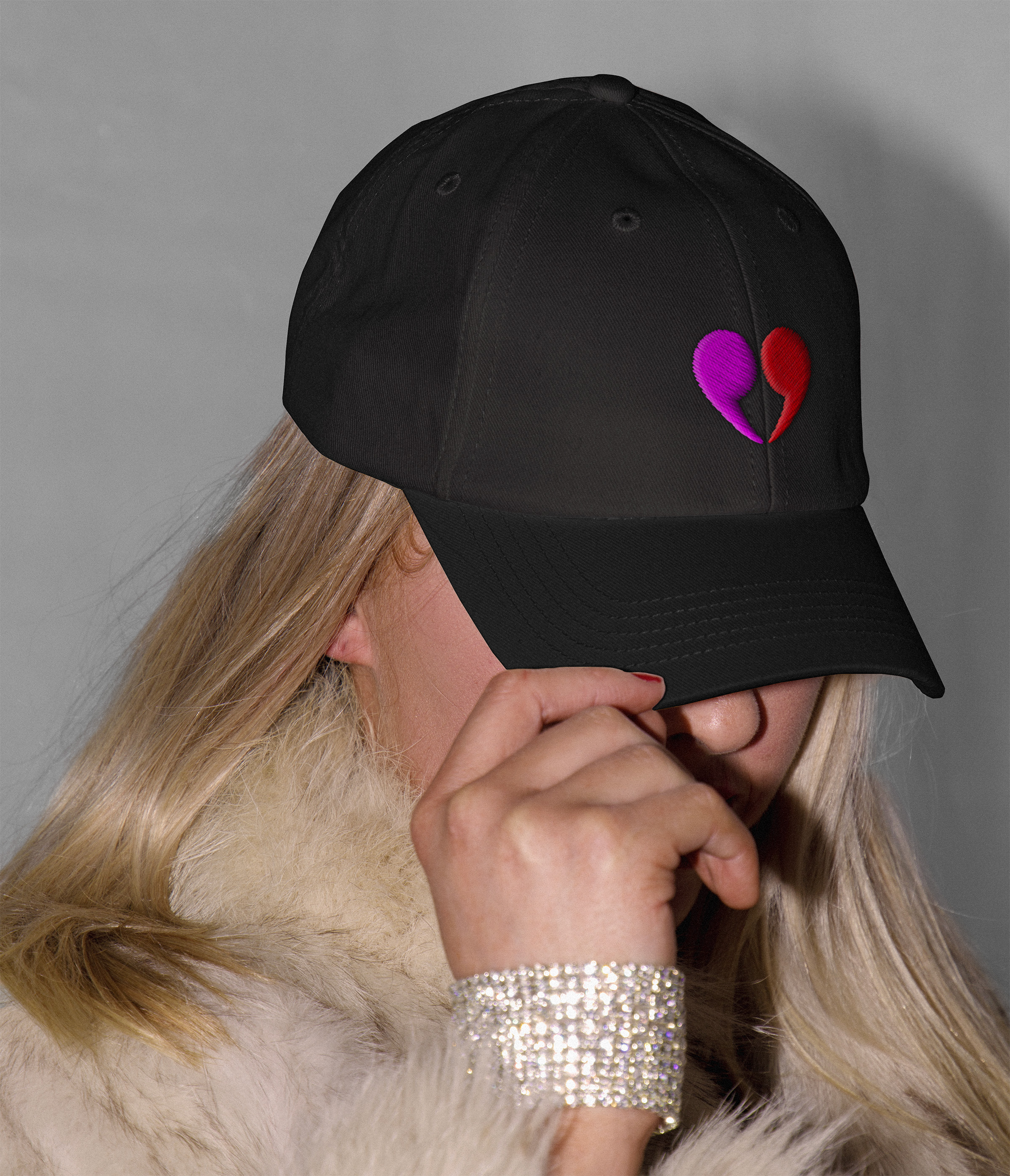 This is an image of a blond woman wearing a black baseball cap with the heart quotation mark symbol embroidered on it. She's tipping it forward so you can't see her face and the symbol is very clear.