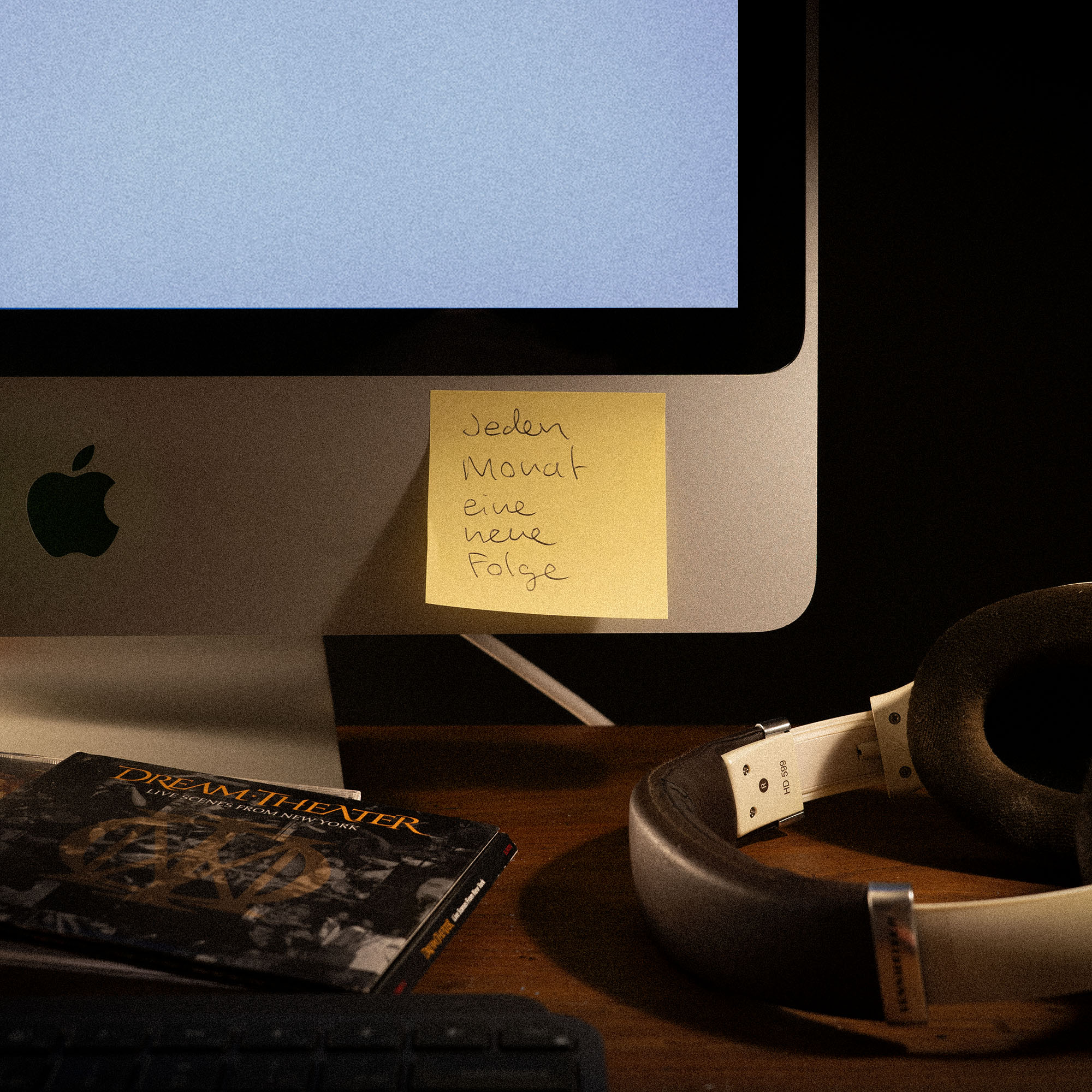 Close-up of an Apple iMac with a yellow sticky note that reads 'Jeden Monat eine neue Folge,' a Dream Theater CD, white headphones, and text announcing a radio show on January 12, 2026 at 22:00.