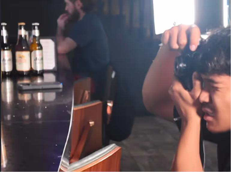 Person holding a camera close to their face, taking a photo in a dimly lit bar with bottles on the counter and another person sitting in the background.