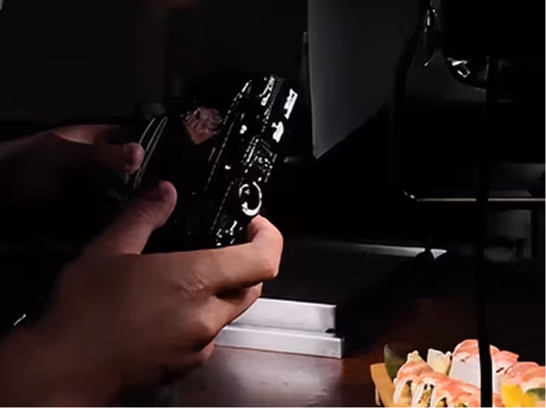 Hands holding a camera focused on sushi rolls under studio lighting on a table.