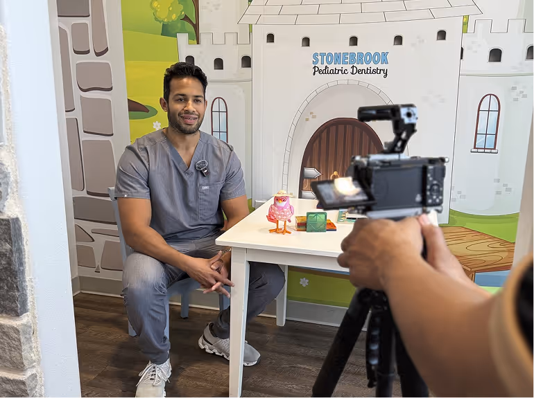 Man in gray scrubs sitting in a pediatric dentistry office with a camera recording him.