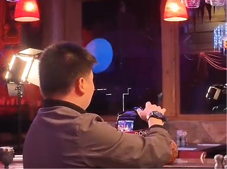 Man with short black hair wearing a brown jacket, adjusting a camera on a tripod in a dimly lit room with red hanging lamps and window reflections.