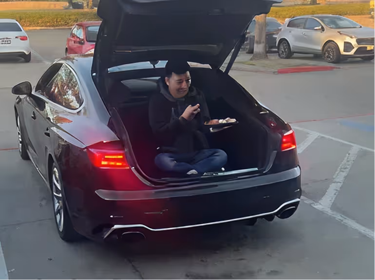 Person sitting cross-legged in the open trunk of a black car, eating food from a plate with chopsticks in a parking lot.