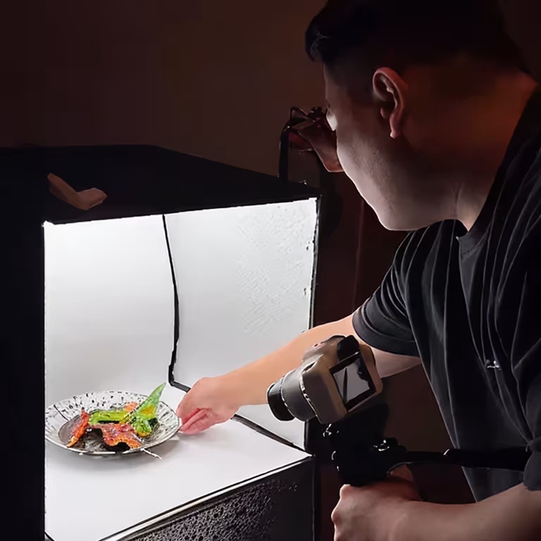 Person adjusting a plate with colorful food inside a lightbox for photography while holding a camera.