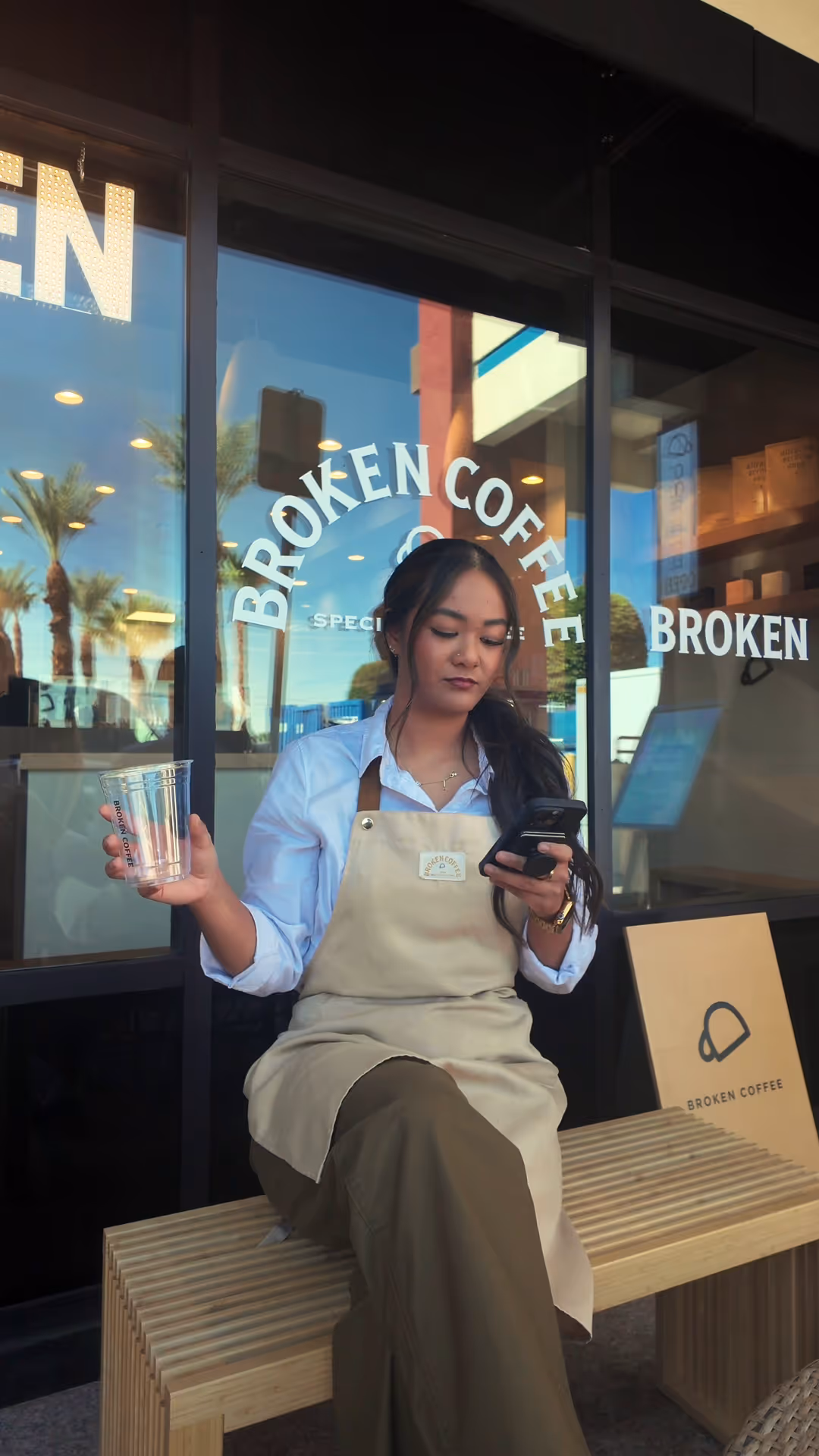 Woman in a beige apron holding a clear plastic cup and looking at her phone while sitting on a wooden bench outside Broken Coffee shop.