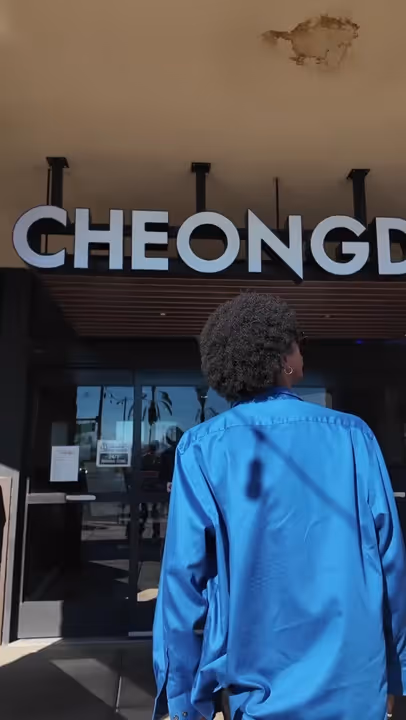 Person with curly hair wearing a blue shirt standing in front of building entrance with large 'CHEONGD' sign overhead.