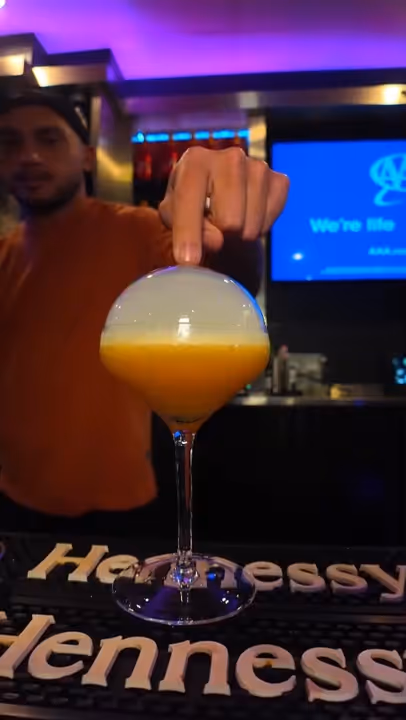 Bartender in an orange shirt touching a cocktail glass with an orange drink and a smoke bubble on top at a bar counter with Hennessy branding.