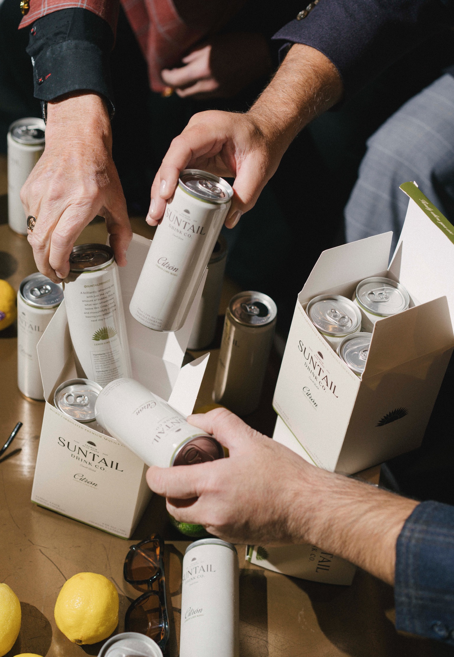 Hand holding a cold Suntail Drink Co. Citron lime margarita can surrounded by lemons, limes, other Suntail cans, and a watermelon slice.