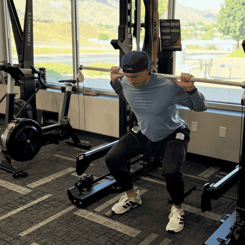 Athlete performing a squat exercise in a gym while wearing blood flow restriction (BFR) cuffs on both thighs. He holds a barbell across his shoulders, demonstrating proper form for low-load strength training with BFR equipment.