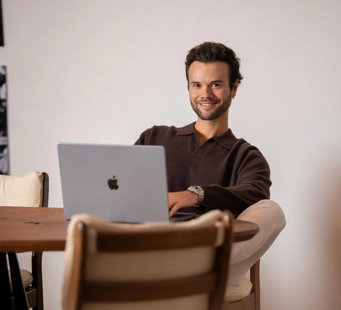 Junger Mann sitzt an einem Holztisch und arbeitet mit einem Apple Laptop, während er lächelnd in die Kamera schaut.