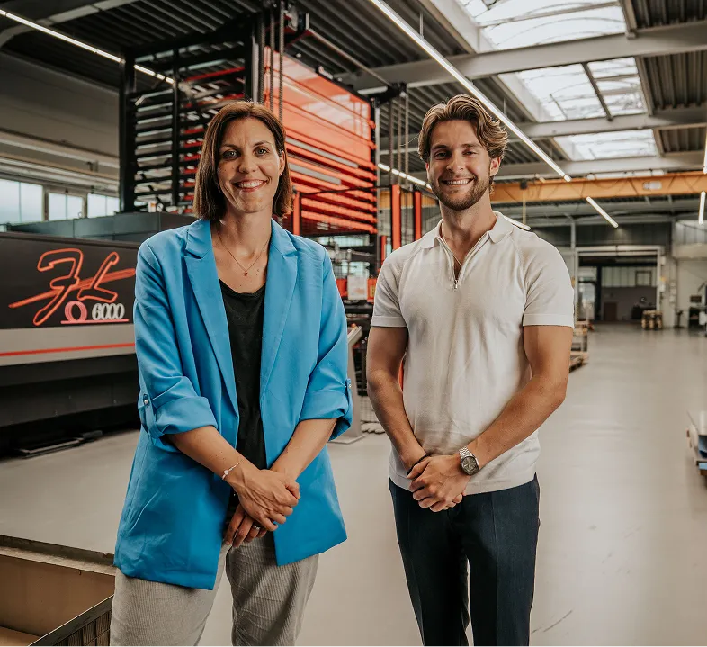 Zwei lächelnde Personen, eine Frau in einem blauen Blazer und ein Mann in weißem Polohemd, stehen in einer modernen Industriehalle.