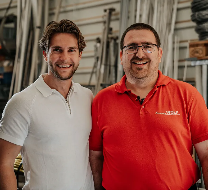 Zwei lächelnde Männer, einer im weißen Polohemd und der andere im roten Polohemd mit Logo, stehen in einer Werkstatt.