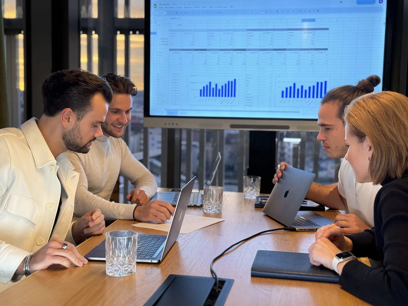 Vier junge Erwachsene arbeiten in einem modernen Besprechungsraum an Laptops mit einer großen Bildschirmpräsentation von Diagrammen im Hintergrund.