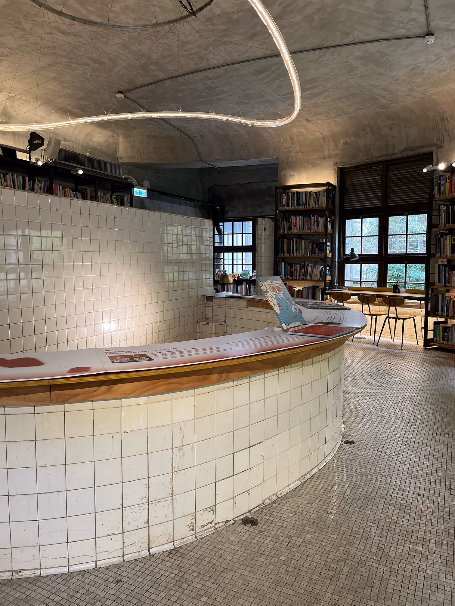 Curved white tiled counter with bookshelves and seating by large window in cozy library space.