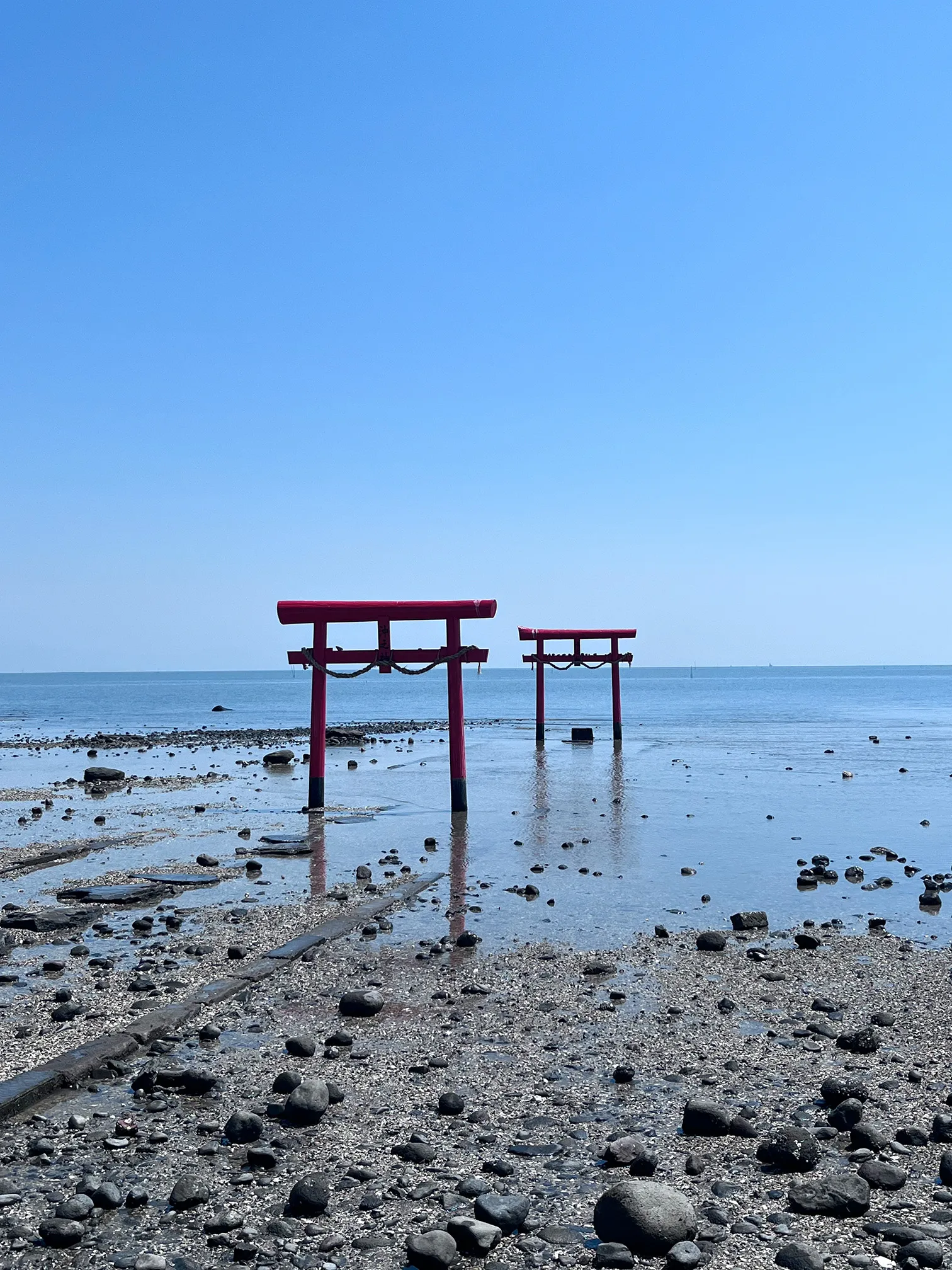 Two red torii gates standing in shallow water near a rocky shore beneath a clear blue sky.