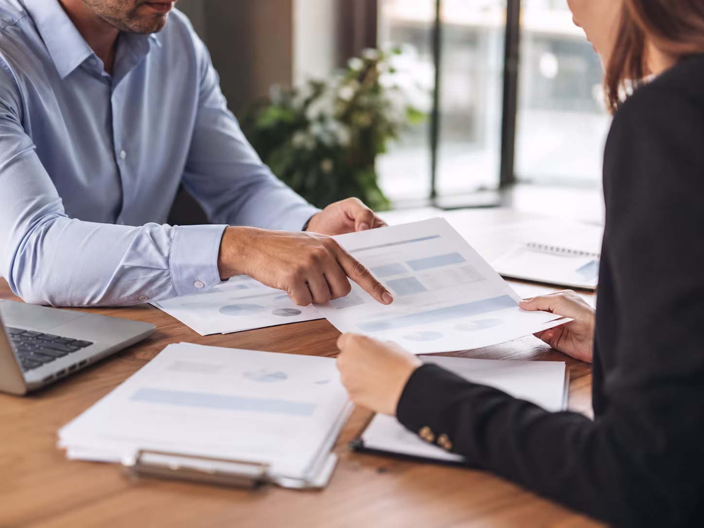 Two business professionals sit across a wooden desk in a modern office, reviewing printed reports together as one points to a document, with a laptop and softly blurred windows in the background.