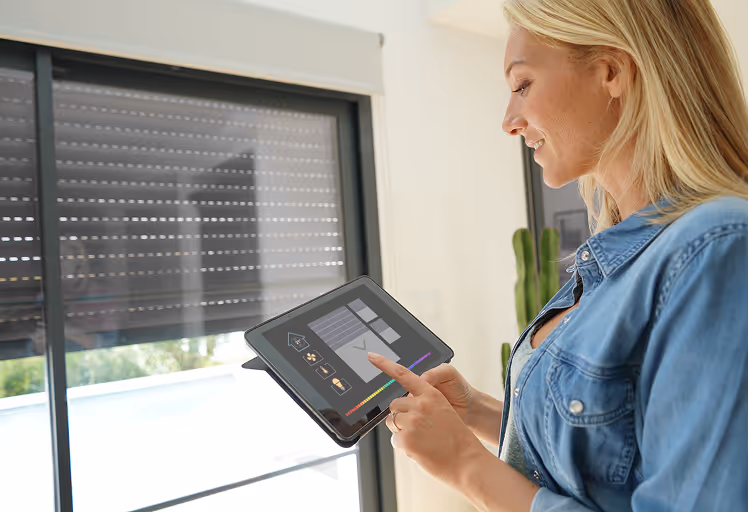 Frau steuert Smart-Home-System an einem Tablet in modernem Wohnzimmer mit großen Fenstern.