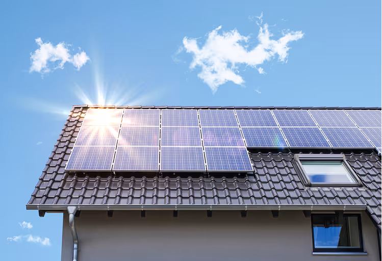 Hausdach mit Solarzellen unter blauem Himmel und Sonnenstrahlen.
