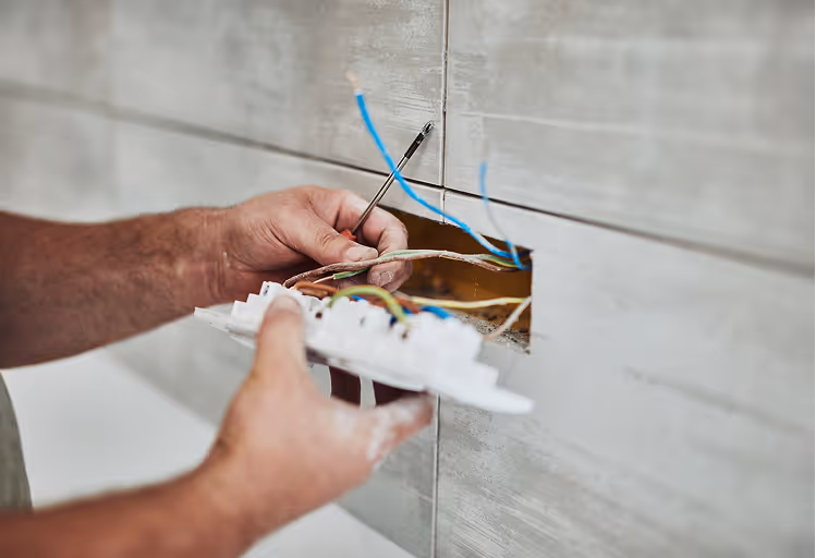 Hände eines Elektrikers, der eine Steckdose mit mehreren Drähten in einer gefliesten Wand installiert.