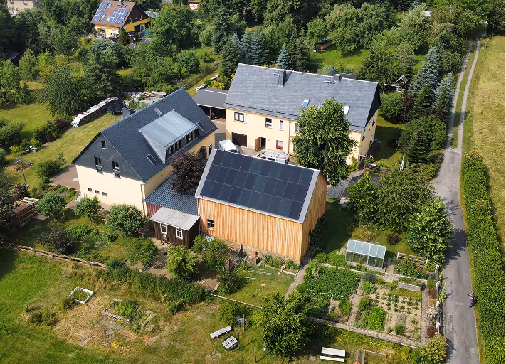 Luftaufnahme eines Wohngebiets mit Häusern, Gärten und Solarpaneelen auf Dächern, umgeben von Bäumen.