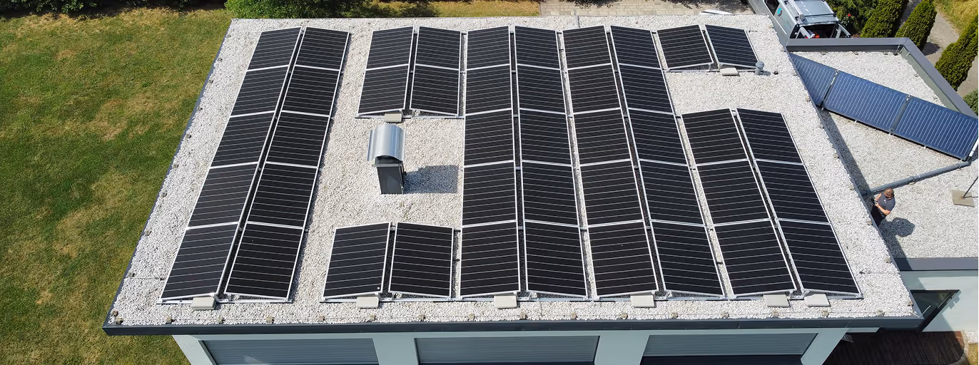 Flachdach eines Gebäudes mit mehreren Solarzellen und einer Belüftungseinheit in der Mitte, umgeben von grünem Rasen.