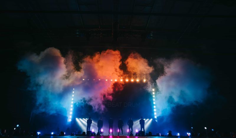 Huge billow of orange and blue smoke rising over the stage as six P1Harmony members stand in light