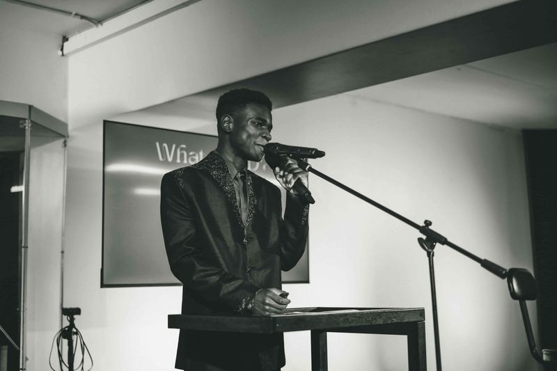 Black and white shot of Josh in a patterned jacket at a lectern with a microphone, a screen behind him