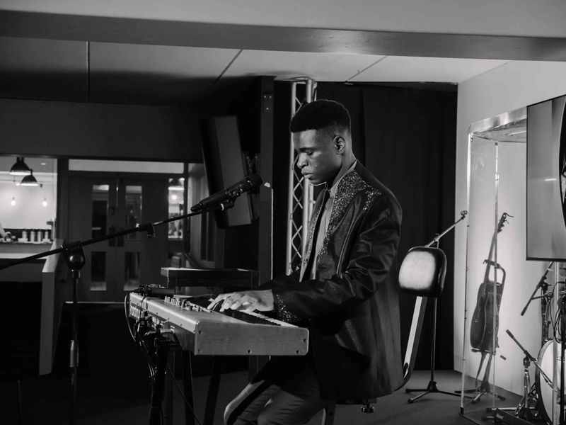 Black and white portrait of Josh in a patterned jacket playing keyboard, guitars on stands behind