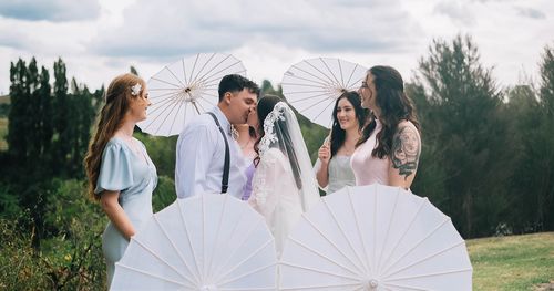 Joe kissing Mia under her veil beside bridesmaids holding white paper parasols