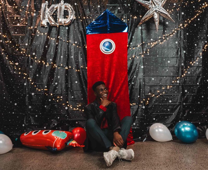 Josh laughing as he sits cross-legged beneath a red rocket prop and silver KD balloon letters