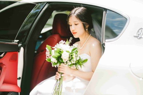 May seated in the open door of a white car with red seats, eyes lowered to her white bouquet