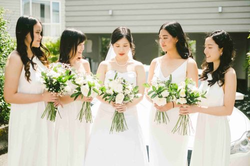 May and her bridesmaids in white in a row, looking down at their bouquets in the midday sun