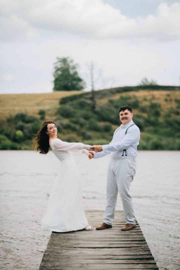 Mia and Joe holding hands and leaning apart on a wooden jetty, green farmland hills behind