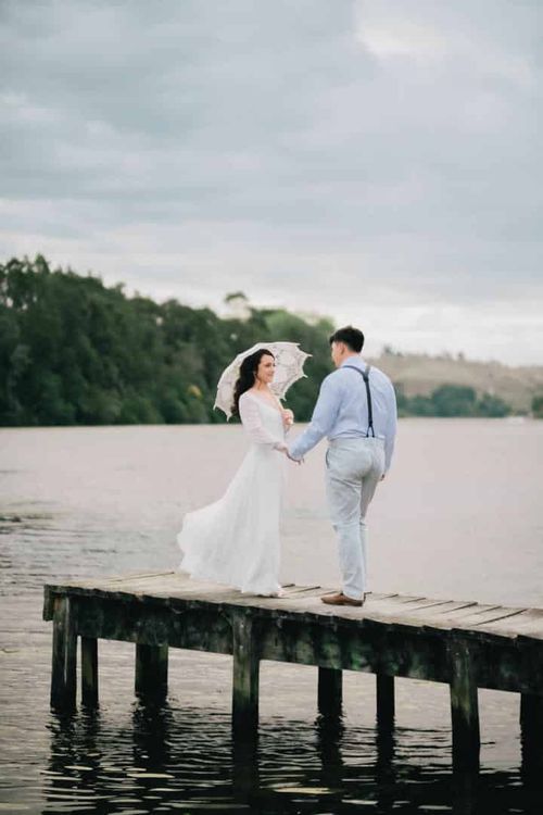 Mia and Joe holding hands on a wooden jetty over the lake, Mia holding a white paper parasol