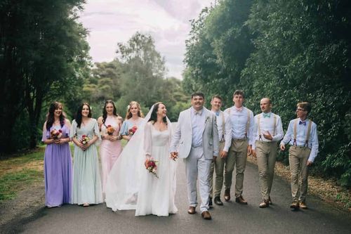 Mia and Joe walking down a tree-lined road with bridesmaids in pastel dresses and groomsmen in braces