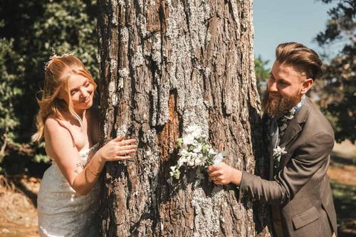Rebekah and Mathew peeking at each other around a pine tree trunk, Mathew holding white flowers