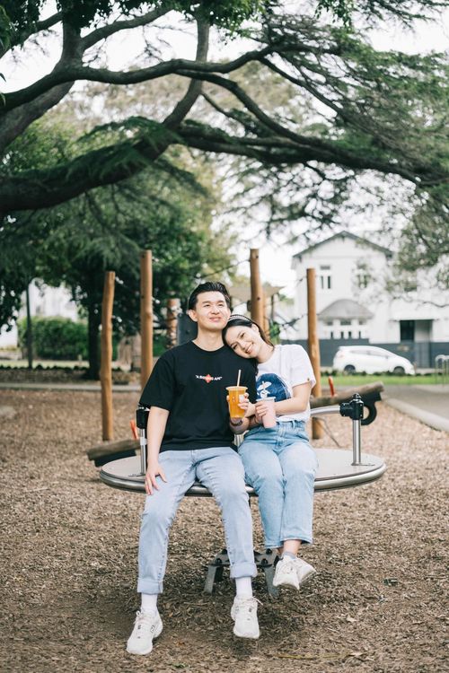 Suzen resting her head on Nick's shoulder on a playground roundabout under spreading branches