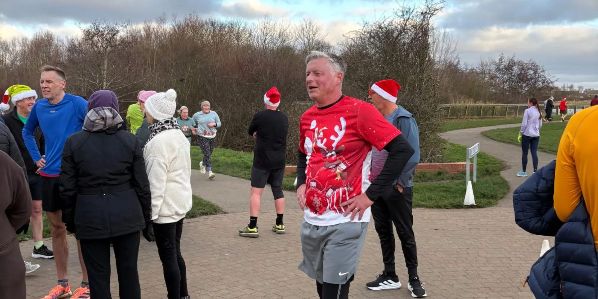 A group of people at a park during a "Santa Dash" event. A man in the center wears a bright red shirt with a large reindeer graphic. Other participants stand around chatting, some wearing Santa hats, with a paved walking path.