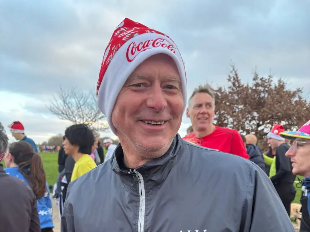 A close-up, smiling portrait of a man wearing a red and white Coca-Cola branded beanie hat. He is standing outdoors in a crowd of people, some of whom are also wearing festive attire, under an overcast sky.