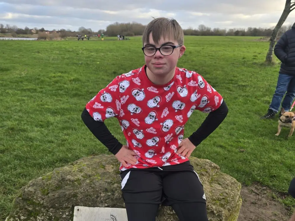 A portrait of a young man wearing glasses and a red t-shirt covered in a repeating Santa Claus pattern. He is sitting on a large rock in an open green field, with his hands on his hips, posing for the camera.