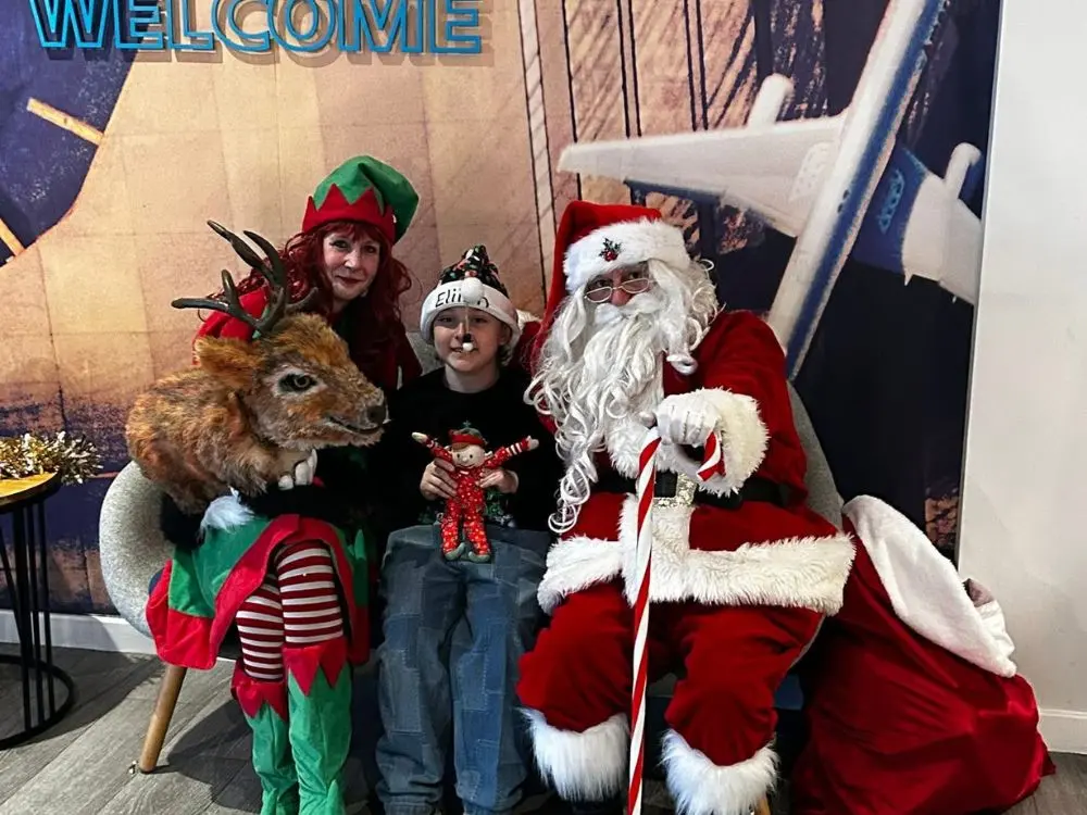 A child holding a small toy poses with Father Christmas and a festive Elf in front of a welcome sign.